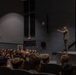 A military officer in camouflage uniform stands on a stage addressing a large audience of uniformed military personnel seated in an auditorium. The speaker is gesturing with one arm extended, appearing to be delivering a briefing or command address. The setting is a formal military theater or auditorium with stage lighting and gold curtains.