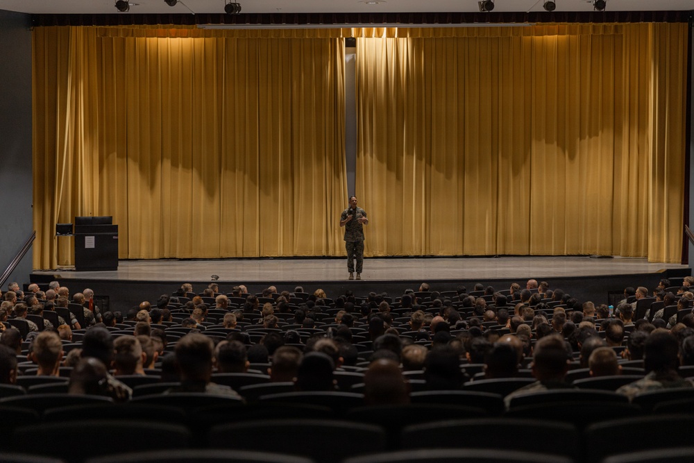 A person in military camouflage uniform stands on a stage in front of large gold/yellow curtains, addressing a large audience seated in an auditorium. The audience appears to consist predominantly of military personnel in uniform, filling most of the visible seating rows. The setting appears to be a formal military briefing, lecture, or all-hands meeting held in a theater-style venue.