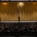 A person in military camouflage uniform stands on a stage in front of large gold/yellow curtains, addressing a large audience seated in an auditorium. The audience appears to consist predominantly of military personnel in uniform, filling most of the visible seating rows. The setting appears to be a formal military briefing, lecture, or all-hands meeting held in a theater-style venue.