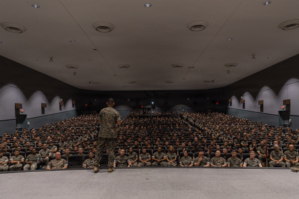 A military officer in camouflage uniform stands on a stage addressing a large auditorium filled with hundreds of uniformed military personnel seated in rows. The setting appears to be a formal military briefing or all-hands meeting held in a large theater-style venue. The audience is attentive and seated in organized rows filling the entire seating area.