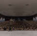 A military officer in camouflage uniform stands on a stage addressing a large auditorium filled with hundreds of uniformed military personnel seated in rows. The setting appears to be a formal military briefing or all-hands meeting held in a large theater-style venue. The audience is attentive and seated in organized rows filling the entire seating area.