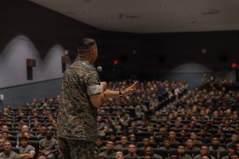 A military officer in camouflage uniform stands on a stage addressing a large auditorium filled with hundreds of uniformed military personnel. The speaker is holding a microphone and gesturing with one hand toward the seated audience. The setting appears to be a formal military briefing or all-hands assembly in a large indoor theater.