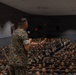 A military officer in camouflage uniform stands on a stage addressing a large auditorium filled with hundreds of uniformed military personnel. The speaker is holding a microphone and gesturing with one hand toward the seated audience. The setting appears to be a formal military briefing or all-hands assembly in a large indoor theater.