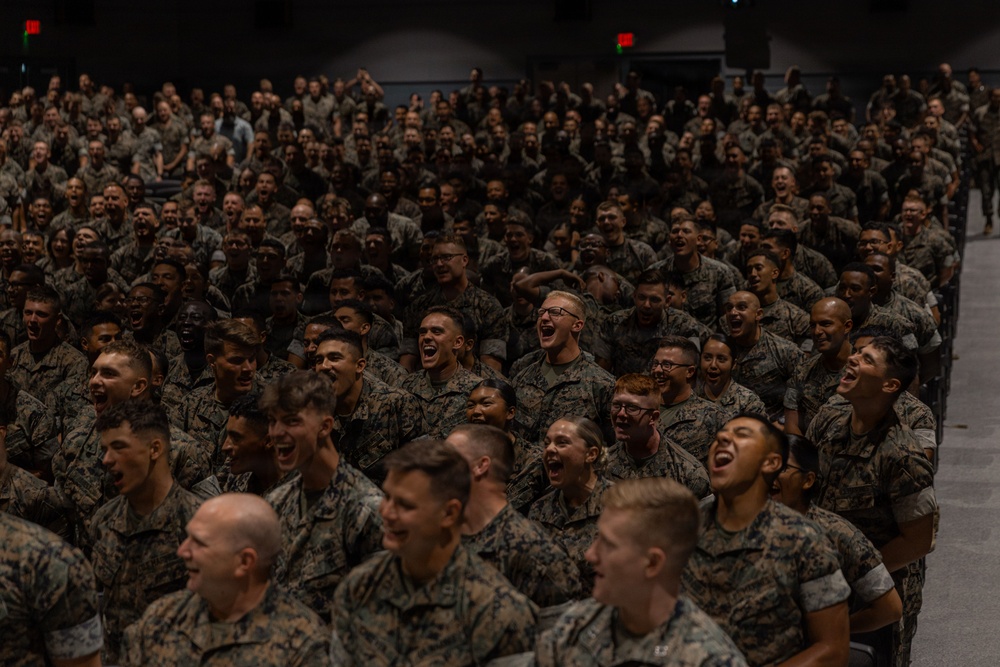 A large assembly of military personnel, all wearing matching camouflage uniforms consistent with U.S. Marine Corps MARPAT pattern, are gathered in what appears to be an auditorium or large indoor venue. The crowd, numbering in the hundreds, appears to be reacting enthusiastically — many individuals have their mouths open as if shouting, cheering, or responding in unison. The scene suggests a formal military gathering such as an all-hands meeting, motivational event, or command presentation.