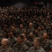 A large assembly of military personnel, all wearing matching camouflage uniforms consistent with U.S. Marine Corps MARPAT pattern, are gathered in what appears to be an auditorium or large indoor venue. The crowd, numbering in the hundreds, appears to be reacting enthusiastically — many individuals have their mouths open as if shouting, cheering, or responding in unison. The scene suggests a formal military gathering such as an all-hands meeting, motivational event, or command presentation.