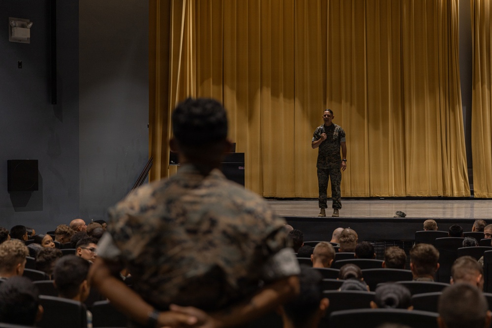 A military briefing or address is taking place in an auditorium or theater setting. A single service member in camouflage uniform stands on a stage in front of a large gold/yellow curtain, speaking to a seated audience of fellow service members. The scene appears to be a formal military assembly or all-hands meeting.