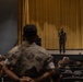 A military briefing or address is taking place in an auditorium or theater setting. A single service member in camouflage uniform stands on a stage in front of a large gold/yellow curtain, speaking to a seated audience of fellow service members. The scene appears to be a formal military assembly or all-hands meeting.