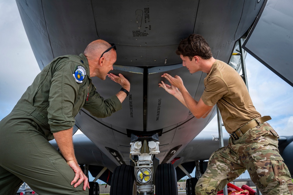 Two military personnel are conducting a close inspection of the underside of a military aircraft, likely during a pre-flight or maintenance check. One individual is wearing an olive green flight suit with a squadron patch, while the other is in camouflage (OCP/MultiCam) uniform. Both are leaning in and pointing at a specific area of the aircraft's fuselage near the nose gear.