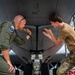 Two military personnel are conducting a close inspection of the underside of a military aircraft, likely during a pre-flight or maintenance check. One individual is wearing an olive green flight suit with a squadron patch, while the other is in camouflage (OCP/MultiCam) uniform. Both are leaning in and pointing at a specific area of the aircraft's fuselage near the nose gear.