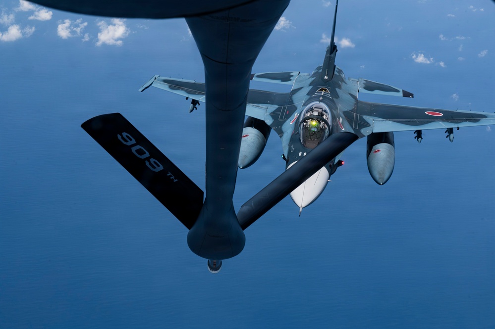 An aerial refueling operation is taking place between a tanker aircraft and a Japan Air Self-Defense Force (JASDF) F-2 fighter jet. The image is taken from the boom operator's position aboard the tanker, looking down at the fighter approaching the refueling boom. The F-2 displays Japanese military markings including the Hinomaru (rising sun) roundel and a two-tone blue/gray camouflage scheme.
