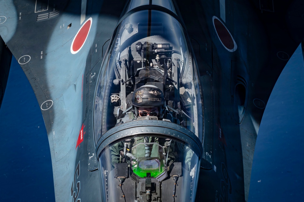 An aerial close-up photograph taken from above showing a military jet fighter pilot in the cockpit during flight. The aircraft displays red roundel markings consistent with the Japan Air Self-Defense Force (JASDF), and the image appears to be taken from a tanker or chase aircraft during an aerial refueling or formation flight. The pilot is wearing a flight helmet with visible markings and sunglasses, with a green HUD (Heads-Up Display) visible in the foreground.