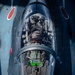 An aerial close-up photograph taken from above showing a military jet fighter pilot in the cockpit during flight. The aircraft displays red roundel markings consistent with the Japan Air Self-Defense Force (JASDF), and the image appears to be taken from a tanker or chase aircraft during an aerial refueling or formation flight. The pilot is wearing a flight helmet with visible markings and sunglasses, with a green HUD (Heads-Up Display) visible in the foreground.