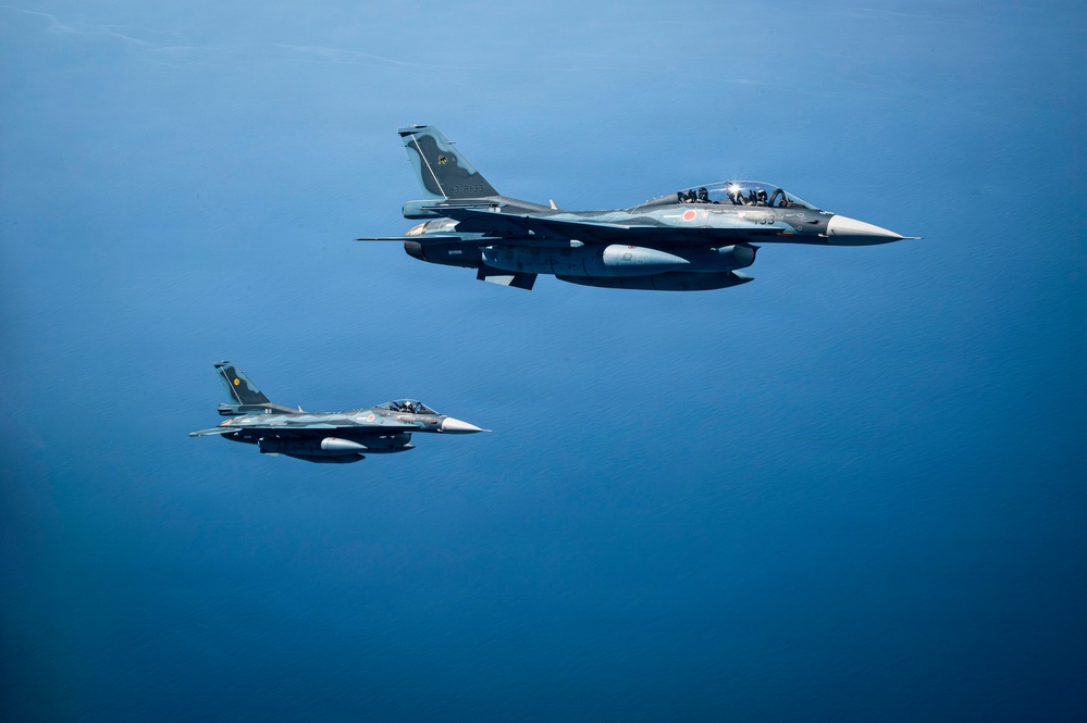 Two military fighter jets are flying in formation over a large body of open blue water. The lead aircraft displays a red roundel marking consistent with the Japan Air Self-Defense Force (JASDF), identifiable as a Mitsubishi F-2 variant. The aircraft are photographed from a close aerial vantage point, likely from a third aircraft, in clear daylight conditions.