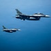 Two military fighter jets are flying in formation over a large body of open blue water. The lead aircraft displays a red roundel marking consistent with the Japan Air Self-Defense Force (JASDF), identifiable as a Mitsubishi F-2 variant. The aircraft are photographed from a close aerial vantage point, likely from a third aircraft, in clear daylight conditions.