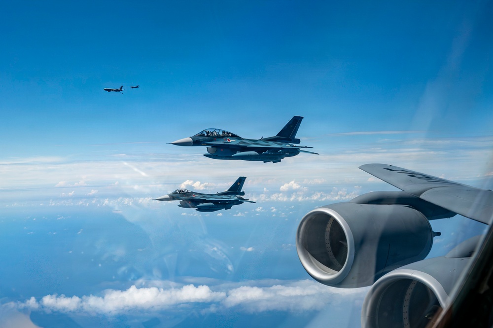 A formation of military aircraft flying at high altitude over a cloud-covered landscape. Two fighter jets bearing Japanese Air Self-Defense Force markings (red circle insignia) fly in close formation alongside what appears to be a large tanker or transport aircraft visible in the foreground. Additional aircraft are visible in the distant background at higher altitude.