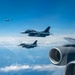 A formation of military aircraft flying at high altitude over a cloud-covered landscape. Two fighter jets bearing Japanese Air Self-Defense Force markings (red circle insignia) fly in close formation alongside what appears to be a large tanker or transport aircraft visible in the foreground. Additional aircraft are visible in the distant background at higher altitude.