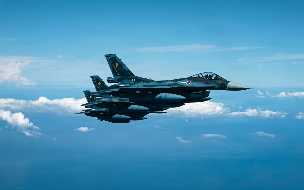 Two military jet fighters are flying in close formation over a body of water with scattered clouds visible in the background. The lead aircraft displays a red circle roundel on its fuselage, consistent with Japan Air Self-Defense Force (JASDF) markings, and carries multiple underwing stores including missiles and fuel tanks. The aircraft appear to be Mitsubishi F-2 fighters, a Japanese-developed variant based on the F-16 design.