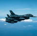 Two military jet fighters are flying in close formation over a body of water with scattered clouds visible in the background. The lead aircraft displays a red circle roundel on its fuselage, consistent with Japan Air Self-Defense Force (JASDF) markings, and carries multiple underwing stores including missiles and fuel tanks. The aircraft appear to be Mitsubishi F-2 fighters, a Japanese-developed variant based on the F-16 design.