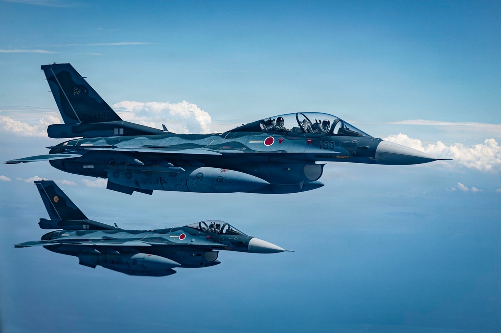 Two Japan Air Self-Defense Force (JASDF) F-2 fighter jets flying in formation over open water or clouds. The lead aircraft bears the tail number '133' and displays the Japanese Hinomaru (red circle) roundel on its fuselage. Both aircraft are painted in a dark blue-gray camouflage scheme typical of JASDF F-2s.