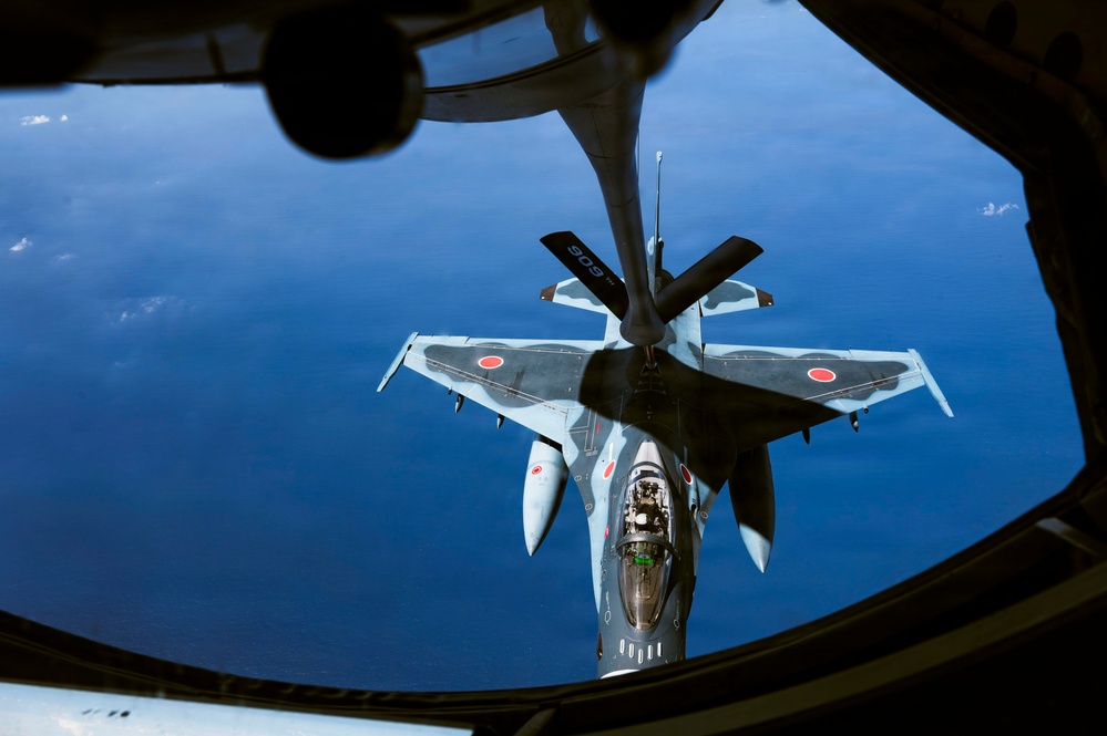An aerial refueling operation is captured from the boom operator's window of a tanker aircraft, showing a Japan Air Self-Defense Force (JASDF) F-2 fighter jet receiving fuel via a flying boom. The F-2 aircraft displays distinctive Japanese roundel markings (red circles) on its wings and fuselage. The deep blue ocean is visible far below, indicating high-altitude flight.