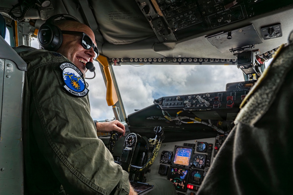A smiling military aviator is seated in the cockpit of what appears to be a large military aircraft, likely a tanker. He is wearing a flight suit with a squadron patch reading 'Air Refueling' and 'Always There,' along with aviation headset and sunglasses. The cockpit displays a complex array of analog and digital instruments, with cloudy skies visible through the windscreen.