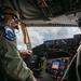 A smiling military aviator is seated in the cockpit of what appears to be a large military aircraft, likely a tanker. He is wearing a flight suit with a squadron patch reading 'Air Refueling' and 'Always There,' along with aviation headset and sunglasses. The cockpit displays a complex array of analog and digital instruments, with cloudy skies visible through the windscreen.