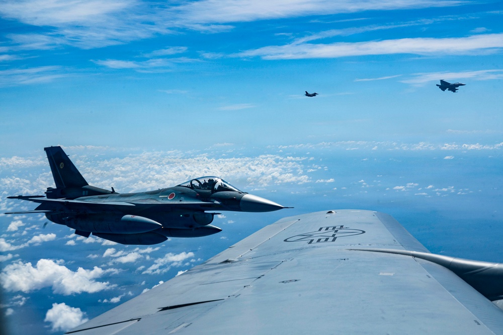 An aerial photograph taken from the fuselage or wing of a large military aircraft, likely a tanker, showing a Japanese Air Self-Defense Force (JASDF) F-2 fighter jet flying in close formation. The F-2 is identifiable by the Japanese hinomaru (red circle) roundel on its tail. Two additional fighter aircraft are visible in the distant background flying in loose formation.