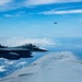 An aerial photograph taken from the fuselage or wing of a large military aircraft, likely a tanker, showing a Japanese Air Self-Defense Force (JASDF) F-2 fighter jet flying in close formation. The F-2 is identifiable by the Japanese hinomaru (red circle) roundel on its tail. Two additional fighter aircraft are visible in the distant background flying in loose formation.