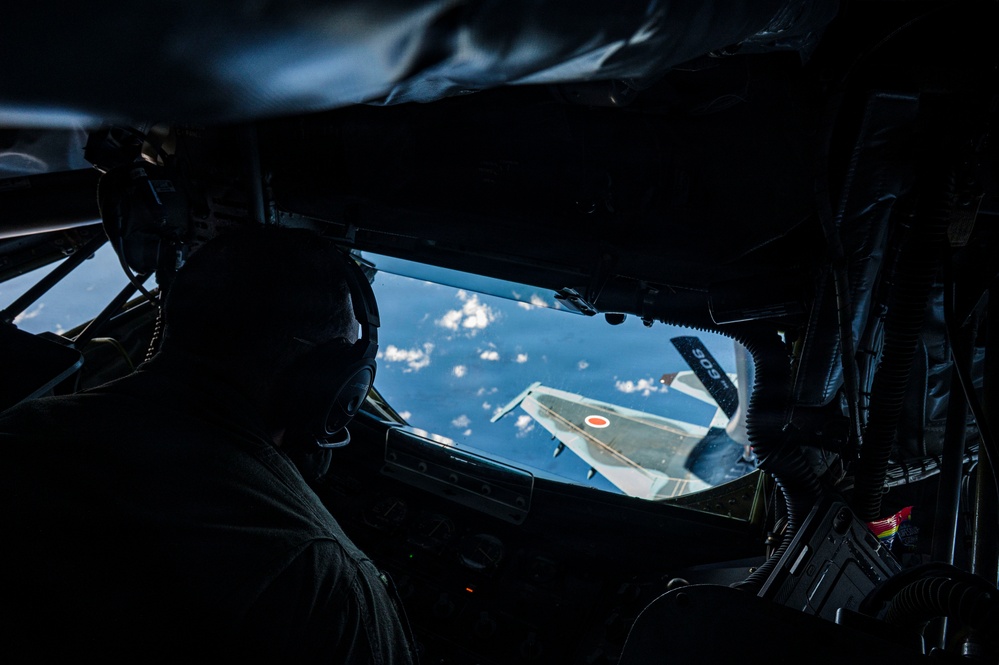 The image shows a view from inside a military tanker aircraft, likely from the boom operator's position, looking out a rear window at a receiving aircraft during an aerial refueling operation. The receiving aircraft displays a red roundel marking consistent with Japan Air Self-Defense Force (JASDF) insignia, with a tail number partially visible as '906'. The interior is heavily shadowed with a silhouetted crew member visible in the foreground.