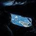 The image shows a view from inside a military tanker aircraft, likely from the boom operator's position, looking out a rear window at a receiving aircraft during an aerial refueling operation. The receiving aircraft displays a red roundel marking consistent with Japan Air Self-Defense Force (JASDF) insignia, with a tail number partially visible as '906'. The interior is heavily shadowed with a silhouetted crew member visible in the foreground.