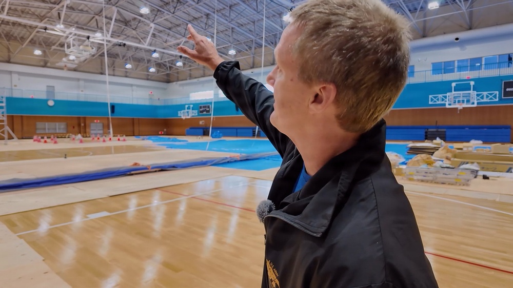 A man in a black jacket is standing inside a large indoor gymnasium, pointing upward toward the ceiling or upper structure. The facility appears to be undergoing renovation or setup, with construction materials and blue floor coverings visible on the hardwood court. The gym features blue accent walls, retractable basketball hoops, and industrial ceiling lighting.