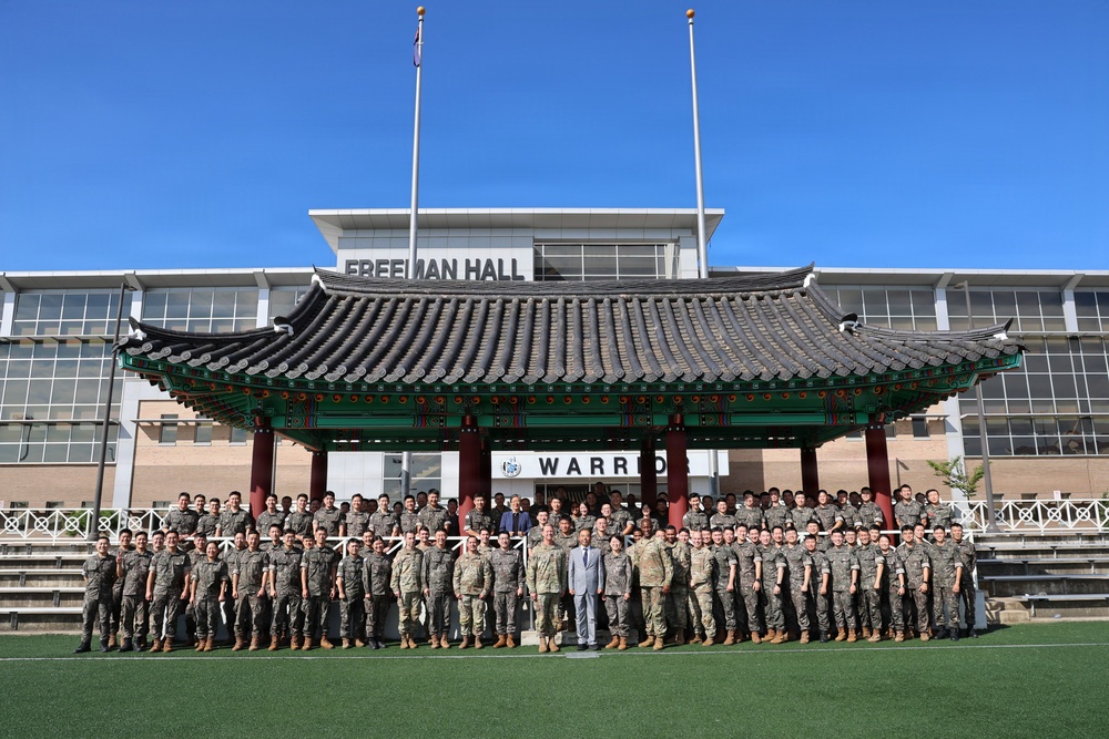 A large group of military personnel, predominantly in camouflage uniforms, pose for a formal group photograph in front of a traditional Korean pavilion (정자/jeongja) structure. The setting appears to be a military installation, with a building labeled 'Freeman Hall' visible in the background along with a partial 'WARRIOR' sign. The combination of the Korean architectural element and mixed military uniforms suggests a joint US-South Korean military event or exchange program.