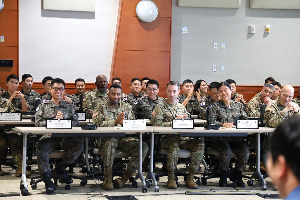 A group of military personnel from what appears to be a joint U.S. Army and Republic of Korea (ROK) Army meeting or briefing session, seated at conference tables in a modern indoor facility. The soldiers are smiling and appear engaged in a positive, collaborative atmosphere. Name placards are visible on the tables, suggesting a formal but friendly exchange or training event.