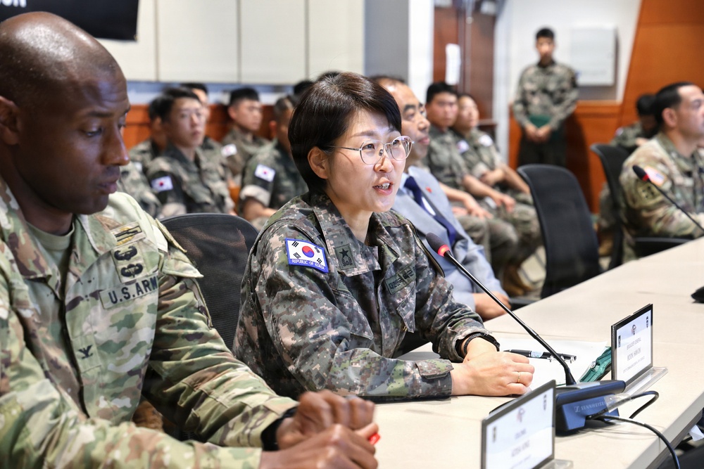 A joint military meeting or briefing is taking place between U.S. Army and Republic of Korea (ROK) Army personnel. A female ROK Army officer wearing a general's star insignia is speaking at a conference table equipped with microphones and name placards. The setting appears to be a formal military conference room with both nations' service members seated in attendance.