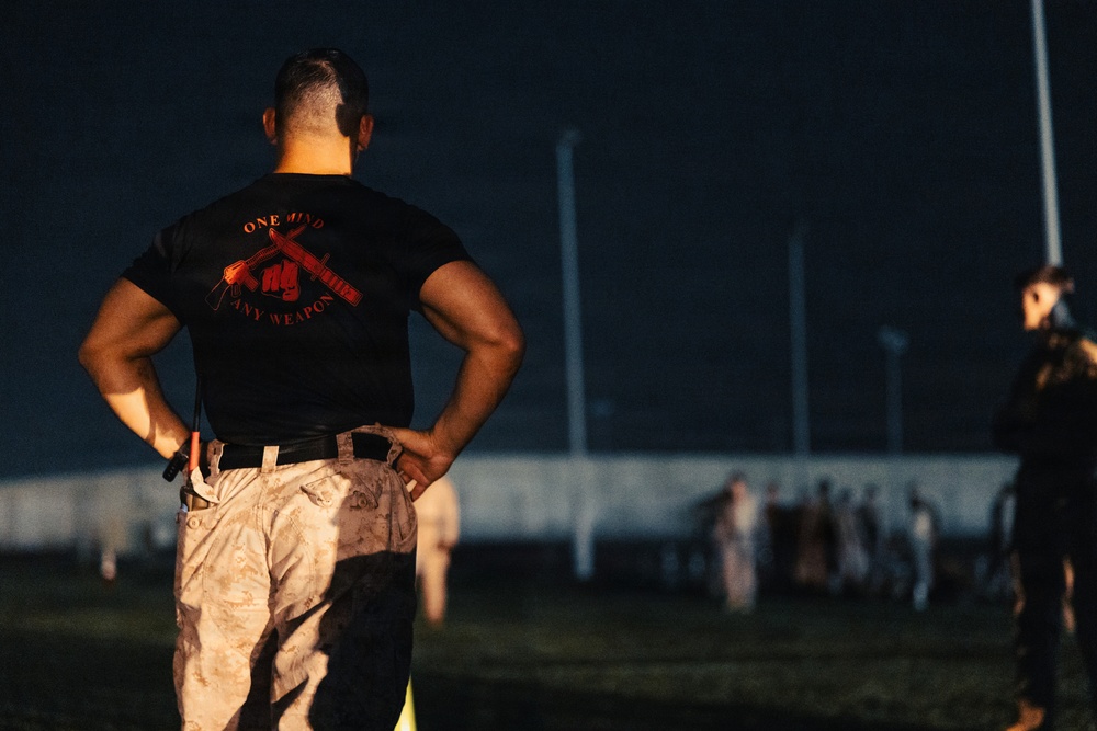 A muscular individual stands with hands on hips, viewed from behind, wearing a black t-shirt reading 'ONE MIND ANY WEAPON' with a crossed-weapons graphic in red. The setting appears to be a military training field at night or low-light conditions. In the background, a group of people in camouflage uniforms can be seen gathered at a distance.