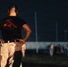 A muscular individual stands with hands on hips, viewed from behind, wearing a black t-shirt reading 'ONE MIND ANY WEAPON' with a crossed-weapons graphic in red. The setting appears to be a military training field at night or low-light conditions. In the background, a group of people in camouflage uniforms can be seen gathered at a distance.