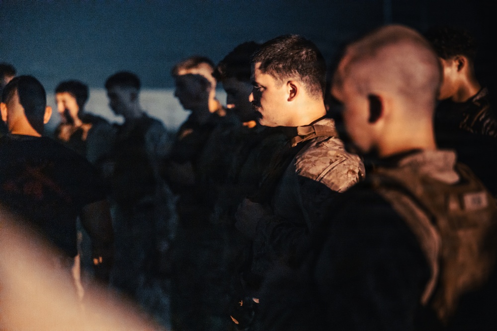 A group of military personnel in uniform are standing in formation during what appears to be a nighttime or low-light setting. The individuals are wearing camouflage and tactical gear, illuminated by warm artificial lighting. The scene suggests a briefing, formation, or pre-mission assembly.