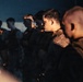 A group of military personnel in uniform are standing in formation during what appears to be a nighttime or low-light setting. The individuals are wearing camouflage and tactical gear, illuminated by warm artificial lighting. The scene suggests a briefing, formation, or pre-mission assembly.