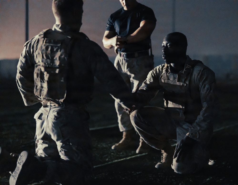 A nighttime military scene showing three personnel in what appears to be a training or briefing situation. One individual in camouflage uniform and tactical vest is crouching, appearing to communicate with standing personnel. The low-light conditions and dramatic lighting create a high-contrast, cinematic appearance.