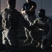 A nighttime military scene showing three personnel in what appears to be a training or briefing situation. One individual in camouflage uniform and tactical vest is crouching, appearing to communicate with standing personnel. The low-light conditions and dramatic lighting create a high-contrast, cinematic appearance.