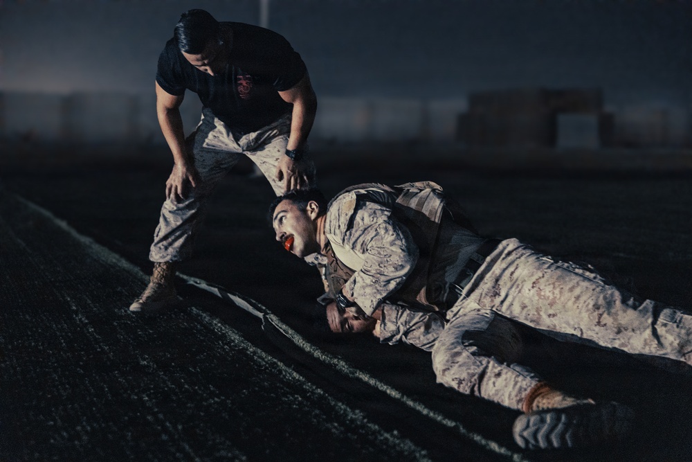 Two individuals in U.S. Marine Corps desert camouflage uniforms (MARPAT) are engaged in what appears to be a combat training or physical fitness drill on a dark outdoor surface, likely a track or field. One person is prone on the ground wearing a tactical vest, while the other stands over them in a bent posture, appearing to assist or instruct. The low-light, dramatic lighting suggests a nighttime training exercise.