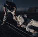Two individuals in U.S. Marine Corps desert camouflage uniforms (MARPAT) are engaged in what appears to be a combat training or physical fitness drill on a dark outdoor surface, likely a track or field. One person is prone on the ground wearing a tactical vest, while the other stands over them in a bent posture, appearing to assist or instruct. The low-light, dramatic lighting suggests a nighttime training exercise.