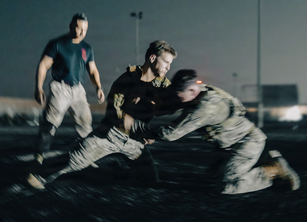 A nighttime scene showing what appears to be a military or law enforcement combatives/hand-to-hand combat training exercise. Two individuals in tactical gear and camouflage uniforms are engaged in a grappling drill, while a third person in a dark t-shirt observes or instructs. The image has significant motion blur, suggesting dynamic movement during a low-light training session.
