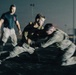 A nighttime scene showing what appears to be a military or law enforcement combatives/hand-to-hand combat training exercise. Two individuals in tactical gear and camouflage uniforms are engaged in a grappling drill, while a third person in a dark t-shirt observes or instructs. The image has significant motion blur, suggesting dynamic movement during a low-light training session.