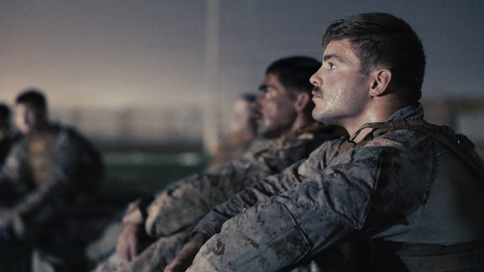 The image shows a group of military personnel in camouflage uniforms seated in a row, appearing to be in a briefing or waiting situation. The foreground subject is a young male soldier with a mustache, wearing a tactical vest over a camouflage uniform, with visible sweat or dirt on his face. The cinematic lighting and composition suggest this may be a still from a film or high-production documentary.