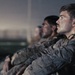 The image shows a group of military personnel in camouflage uniforms seated in a row, appearing to be in a briefing or waiting situation. The foreground subject is a young male soldier with a mustache, wearing a tactical vest over a camouflage uniform, with visible sweat or dirt on his face. The cinematic lighting and composition suggest this may be a still from a film or high-production documentary.