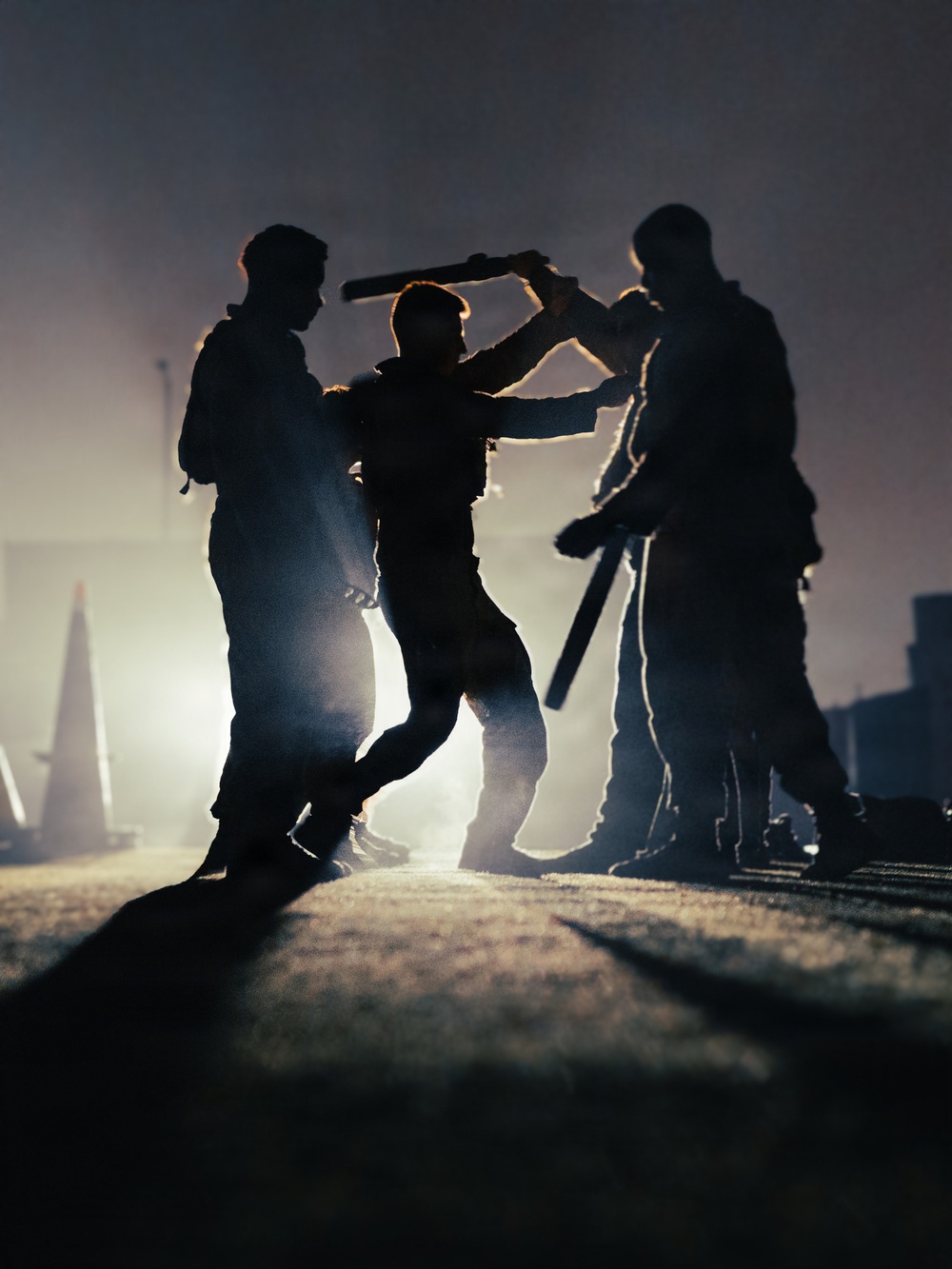 A dramatic backlit silhouette photograph showing three figures engaged in a physical altercation or combat training scenario. The strong backlighting creates stark silhouettes against a hazy, glowing background. At least one figure is wielding a baton or rod-like object in an overhead striking position.