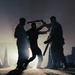 A dramatic backlit silhouette photograph showing three figures engaged in a physical altercation or combat training scenario. The strong backlighting creates stark silhouettes against a hazy, glowing background. At least one figure is wielding a baton or rod-like object in an overhead striking position.