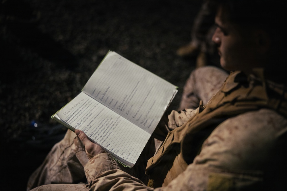 A military personnel in desert camouflage (MARPAT pattern) uniform is reviewing a handwritten notebook in low-light conditions. The notebook contains handwritten notes organized in columns and rows, suggesting operational or field documentation. A second individual in similar uniform is partially visible in the background.