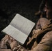A military personnel in desert camouflage (MARPAT pattern) uniform is reviewing a handwritten notebook in low-light conditions. The notebook contains handwritten notes organized in columns and rows, suggesting operational or field documentation. A second individual in similar uniform is partially visible in the background.